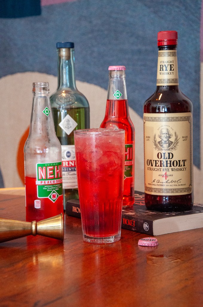 A glass filled with a red drink and ice, surrounded by various liquor bottles including whiskey and absinthe. Some books and a bottle cap are also visible on the wooden table.