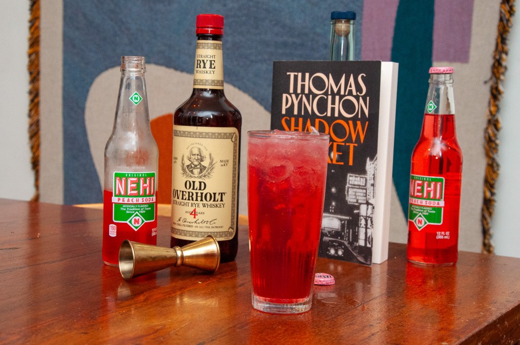 A wooden table featuring a tall glass of red beverage with ice, a bottle of Old Overholt Rye Whiskey, two bottles of Nehi Peach Soda, a jigger, and a book titled 'Thomas Pynchon Shadow Market.' The background includes a textured fabric.